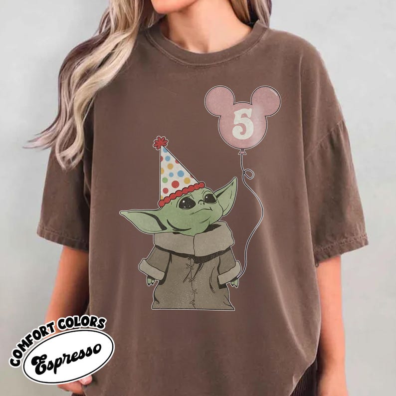 May include: Brown t-shirt featuring a graphic of a green alien-like character wearing a party hat and holding a pink balloon with the number 5. The shirt has the words "COMFORT COLORS Espresso" printed on it.