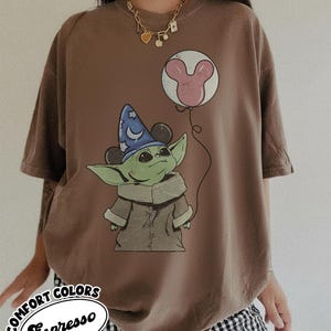 Puede incluir: Camiseta marr&oacute;n con un gr&aacute;fico de un personaje alien&iacute;gena verde con un sombrero de mago y un globo de Mickey Mouse. La camiseta tiene el texto "Comfort Colors Espresso" en un dise&ntilde;o circular.