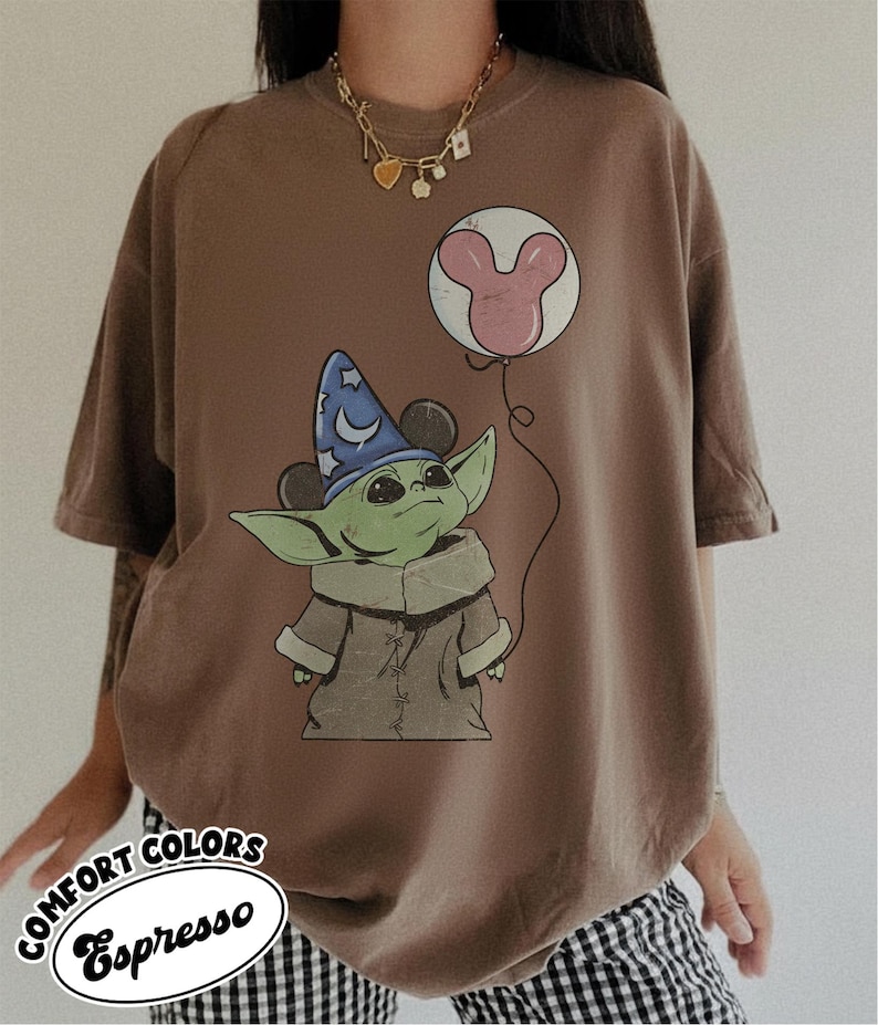 Puede incluir: Camiseta marr&oacute;n con un gr&aacute;fico de un personaje alien&iacute;gena verde con un sombrero de mago y un globo de Mickey Mouse. La camiseta tiene el texto "Comfort Colors Espresso" en un dise&ntilde;o circular.