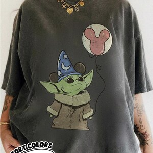 Puede incluir: Camiseta gris oscuro con un personaje verde que lleva un sombrero de mago azul con orejas de Mickey Mouse. El personaje sostiene un globo de Mickey Mouse. La camiseta tiene el texto "COMFORT COLORS Pepper" en un &oacute;valo blanco.
