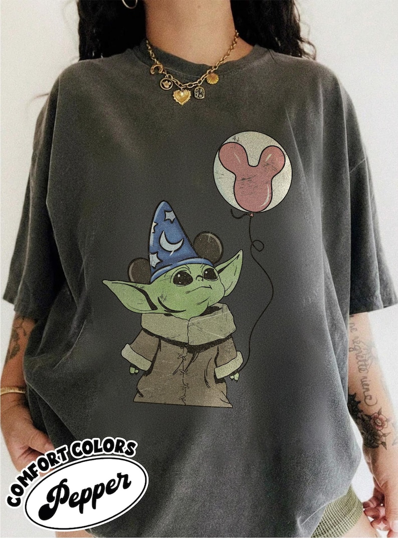 Puede incluir: Camiseta gris oscuro con un personaje verde que lleva un sombrero de mago azul con orejas de Mickey Mouse. El personaje sostiene un globo de Mickey Mouse. La camiseta tiene el texto "COMFORT COLORS Pepper" en un &oacute;valo blanco.