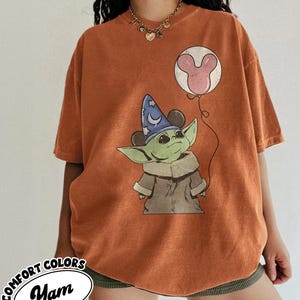 Puede incluir: Camiseta naranja Comfort Colors con un gr&aacute;fico de un personaje alien&iacute;gena verde con un sombrero de mago y un globo de Mickey Mouse. La camiseta tiene el texto "Yam" en un &oacute;valo blanco.