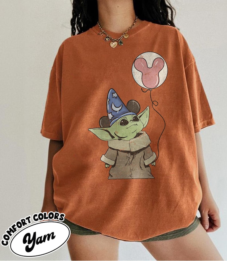 Puede incluir: Camiseta naranja Comfort Colors con un gr&aacute;fico de un personaje alien&iacute;gena verde con un sombrero de mago y un globo de Mickey Mouse. La camiseta tiene el texto "Yam" en un &oacute;valo blanco.