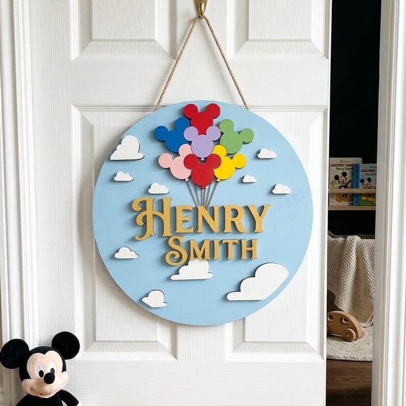 May include: A round, light blue wooden door sign with the name "Henry Smith" in gold lettering. The sign features colorful Mickey Mouse-shaped balloons and white cloud cutouts. A rope is attached for hanging.