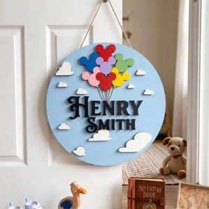 May include: A round, light blue wooden sign with the name "Henry Smith" in black script. The sign features white clouds and colorful balloon-shaped designs. A natural twine is used for hanging.