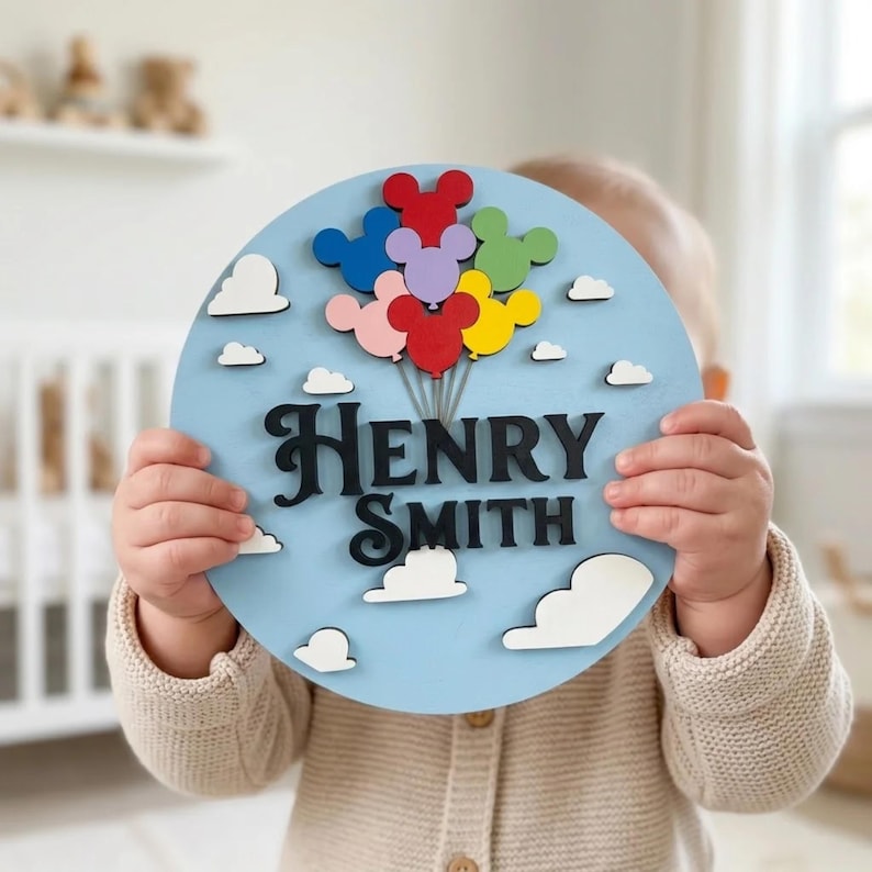 May include: A round, light blue wooden sign with the name "HENRY SMITH" in black, stylized letters. The sign features colorful Mickey Mouse-shaped balloons and white cloud cutouts. A child is holding the sign.