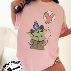 Puede incluir: Camiseta rosa p&aacute;lido Comfort Colors con un personaje de dibujos animados que lleva un sombrero de mago y sostiene un globo con forma de cabeza de Mickey Mouse. La camiseta tiene la inscripci&oacute;n "Comfort Colors Blossom".