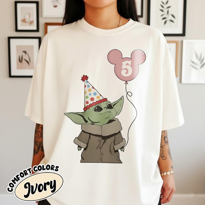 May include: Ivory-colored t-shirt featuring a cartoon character wearing a party hat and holding a pink balloon with the number 5. The t-shirt has the words "COMFORT COLORS" and "Ivory" printed on it.