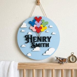 May include: A round, light blue wooden sign with the name "Henry Smith" in black script. The sign features colorful Mickey Mouse-shaped balloons and white cloud cutouts. A wooden airplane and compass are on a shelf below.