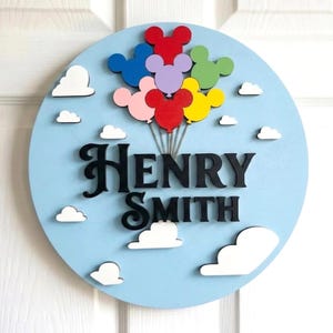 May include: A round, light blue wooden sign with the name "HENRY SMITH" in black script. Colorful Mickey Mouse-shaped balloons float above the name, and white cloud cutouts are scattered around the sign.