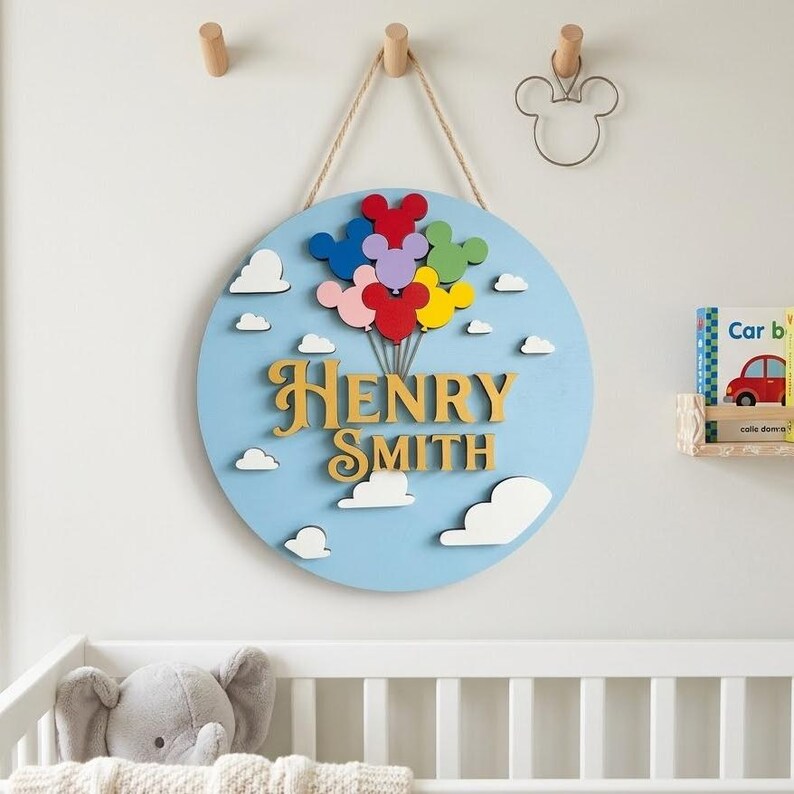 May include: A round, light blue wooden sign with the name "Henry Smith" in gold lettering. The sign features colorful Mickey Mouse-shaped balloons and white cloud cutouts. A rope is used for hanging.