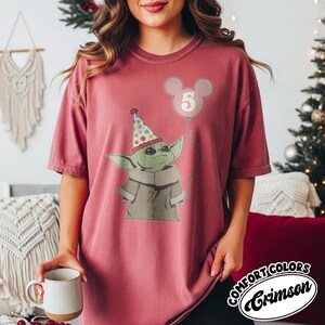 May include: Crimson-colored t-shirt featuring a graphic of a green alien-like character wearing a party hat and holding a balloon with the number 5. The shirt has the words "COMFORT COLORS" and "Crimson" in a circular design.
