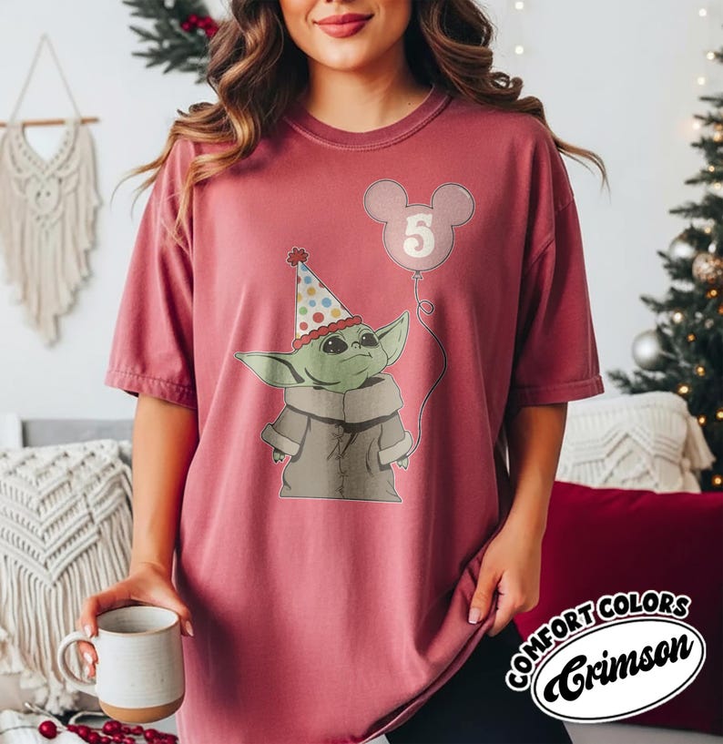 May include: Crimson-colored t-shirt featuring a graphic of a green alien-like character wearing a party hat and holding a balloon with the number 5. The shirt has the words "COMFORT COLORS" and "Crimson" in a circular design.