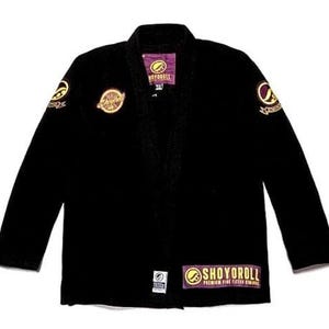 Puede incluir: Uniforme negro de Jiu-Jitsu con detalles dorados y morados. El uniforme presenta parches bordados en las mangas y el pecho, y un parche rectangular con el texto "SHOYOROLL" en la parte inferior. Fondo blanco.