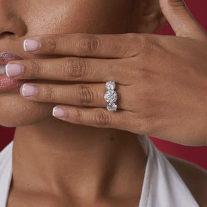 May include: A three-stone diamond ring with large, round-cut diamonds. The ring is set on a finger, with a hand partially covering the face. The background is a solid red color.