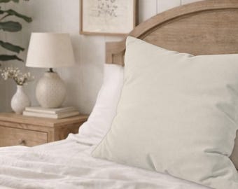 May include: A cream-colored square pillow on a bed with a wooden headboard. The pillow is the focal point, with a soft, textured appearance. A bedside table with a lamp and decorative items is visible in the background.