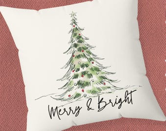 May include: A white square pillow with a hand-drawn Christmas tree design. The tree is green with red ornaments and a star on top. The words "Merry & Bright" are written in black script below the tree. The pillow is on a red textured surface.