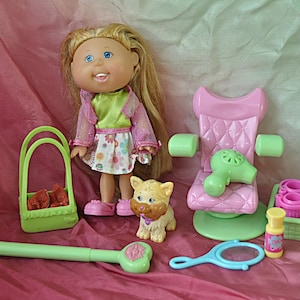 May include: A doll with blonde hair and blue eyes stands next to a pink salon chair and accessories. The set includes a toy dog, a green basket, a hair dryer, a mirror, and other beauty items.
