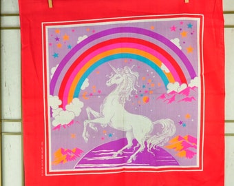 May include: A red bandana with a purple background featuring a white unicorn standing on a purple and orange mountain range. A rainbow with pink, orange, yellow, green, blue, and turquoise stripes arches over the unicorn. The bandana is decorated with white stars.