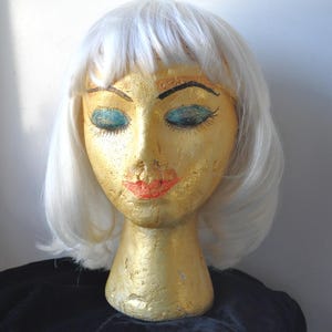 May include: A vintage mannequin head with a gold painted face, closed teal eyes, and red lips. The mannequin is wearing a short, white wig. The head is displayed on a black surface.