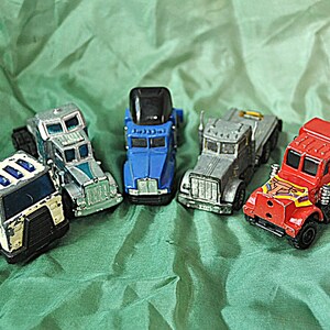 May include: A collection of toy semi-trucks in various colors, including red, blue, silver, and white. The trucks are arranged on a green fabric surface. Each truck has detailed features, such as headlights, grills, and wheels.