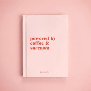 May include: A light pink hardcover book with the text "powered by coffee & sarcasm" printed in a bold red serif font on the front cover. The name "EMILY CARTER" is printed in a smaller, simple red font near the bottom of the book cover.