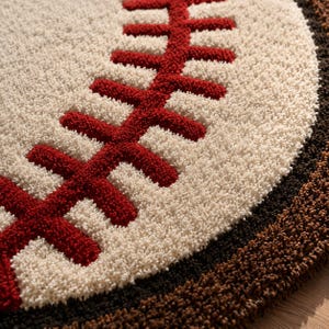 Puede incluir: Una alfombra circular con mechones que presenta un fondo de color crema con un prominente patr&oacute;n de b&eacute;isbol cosido en rojo en el centro. El borde exterior de la alfombra est&aacute; bordeado por una banda gruesa y texturizada de color marr&oacute;n oscuro.