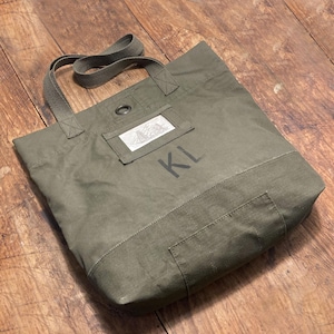 Puede incluir: Bolso tote verde oliva con dos asas y un parche rectangular. El bolso presenta las iniciales "KL" impresas en negro. El bolso está hecho de una tela duradera y tiene forma rectangular.