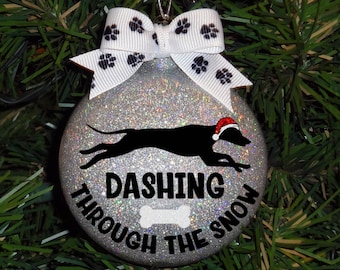 Pode incluir: Um enfeite de Natal de glitter prateado com uma silhueta preta de um cão usando um chapéu de Papai Noel correndo e o texto "Dashing Through The Snow" com um gráfico de osso.