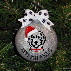 Pode incluir: Enfeite de Natal prateado brilhante com um cão doodle preto e branco usando um gorro de Papai Noel. O enfeite tem uma fita branca com pegadas pretas e o texto "HAPPY HOLI-DOODLES".