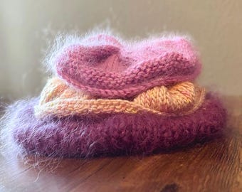 May include: A stack of three knitted items in shades of pink and peach. The bottom item is a fuzzy, deep purple. The middle item is a peach color, and the top item is a knitted pink hat. The items are stacked on a wooden surface.