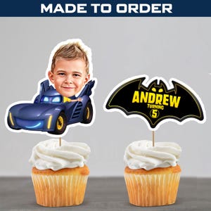 May include: Two cupcakes with white icing, each with a personalised cake topper. One shows a boy in a blue car, the other a black bat symbol with the name "Andrew" and the number "5". The words "MADE TO ORDER" are at the top.