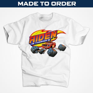Puede incluir: Camiseta blanca con un gráfico colorido de un monster truck, el nombre "AIDEN" y la frase "IS TURNING 3". El diseño incluye llamas y un óvalo amarillo. Las palabras "MADE TO ORDER" están en la parte superior.