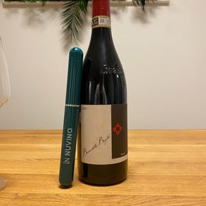 May include: A dark wine bottle with a cork and a label featuring text and a red emblem. A green wine preserver with the text "N NUVINO" is next to the bottle. The bottle is on a wooden surface.