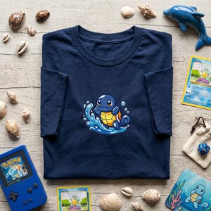 May include: A navy blue t-shirt with an embroidered Squirtle design, depicting the character in blue water waves. The design includes a yellow and orange shell. The image also shows seashells, a blue dolphin ornament, and Pokémon cards.