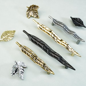 May include: An assortment of leaf-shaped cabinet hardware in various metallic finishes. The image showcases several drawer pulls and knobs in gold, silver, black, and bronze tones. The hardware is designed to resemble leaves, adding a natural aesthetic.