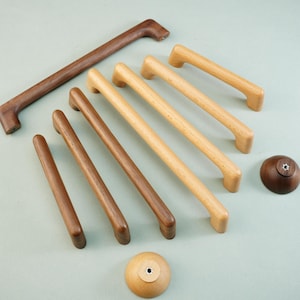 May include: An assortment of wooden cabinet hardware, including handles and knobs, in various shades of brown and natural wood tones. The handles are rectangular and curved, while the knobs are round. The hardware is arranged on a light green surface.