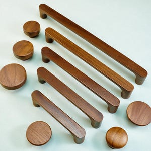 May include: A collection of wooden cabinet hardware, including several sizes of rectangular pulls and round knobs. The hardware is a warm brown color and appears to be made of solid wood. The knobs and pulls are arranged on a light background.