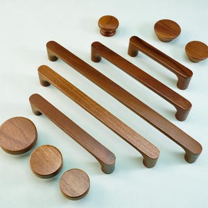 Pode incluir: Uma coleção de ferragens de madeira para armários, incluindo puxadores e botões de vários tamanhos. Os puxadores são retangulares com pegas curvas e os botões são redondos. A ferragem é de cor castanha quente e disposta sobre um fundo claro.