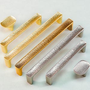 Puede incluir: Una selección de tiradores y pomos para armarios en acabados dorados, bronce y plateados. El herraje presenta una textura martillada. Los tiradores y pomos se muestran sobre un fondo claro. Medidas en centímetros.