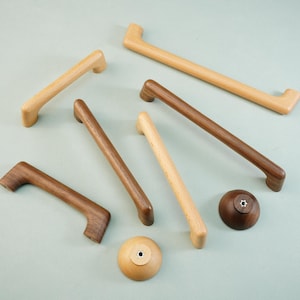 May include: A selection of wooden cabinet handles and knobs in various shades of brown and natural wood. The handles are different lengths, with a curved design. The knobs are round. The hardware is displayed on a pale blue-green surface. Measurements are in cm.