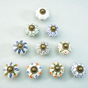 May include: A collection of ten ceramic cabinet knobs with floral designs. The knobs are white with various hand-painted patterns in green, blue, orange, and black. Each knob has a brass-colored metal center and a flower-like shape.
