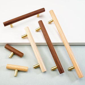 Pode incluir: Uma coleção de puxadores de armário de madeira em vários tons de castanho e madeira natural, cada um com uma pega de cor latão. Os puxadores estão dispostos numa superfície branca, mostrando os seus diferentes tamanhos e grãos de madeira. Medidas em cm.