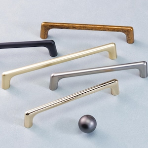 Op de afbeelding: Een selectie van kastgrepen en een knop in verschillende metallic afwerkingen: zwart, goud, zilver en brons. De stanggrepen en de knop worden gepresenteerd op een lichtblauwe ondergrond.