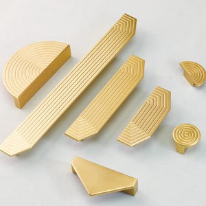May include: A collection of gold-colored cabinet hardware, including handles and knobs. The hardware features geometric designs with linear and semi-circular patterns. The pieces vary in size and shape, suitable for drawers and cabinets.
