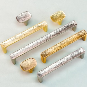 May include: An assortment of cabinet hardware in gold and silver tones. The collection includes bar pulls and oval knobs with a hammered texture. The hardware is displayed on a light blue surface, showcasing the different finishes and styles.