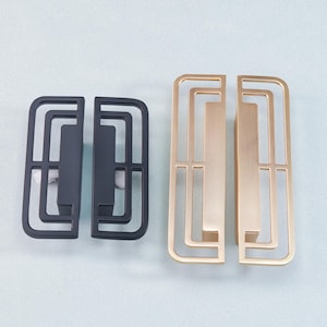 May include: Two pairs of decorative cabinet pulls. One pair is matte black, the other is gold. Each pull has a rectangular shape with a geometric open design. The pulls are made of metal and are suitable for furniture.