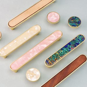 May include: A selection of cabinet hardware, including drawer pulls and knobs. The pulls have rectangular designs with wood, pink, and green accents, while the knobs are round with similar materials. All have gold-toned bases.