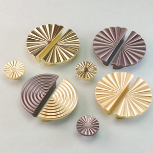 May include: A selection of decorative cabinet hardware in gold and bronze hues. The hardware features round knobs and split-circle pulls with radial and concentric patterns. The hardware is displayed on a light grey surface.