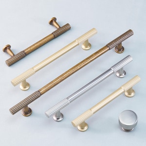 May include: A collection of cabinet hardware, including several bar pulls and a knob. The pulls feature a ribbed design on the bar and are available in various finishes, including brass, silver, and bronze. The knob has a matching ribbed design.